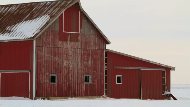 A red barn and the machine shed built onto it, standing in snow under a pale winter sky — the kind of farm outbuilding radiant floor heating is planned for before a slab is poured