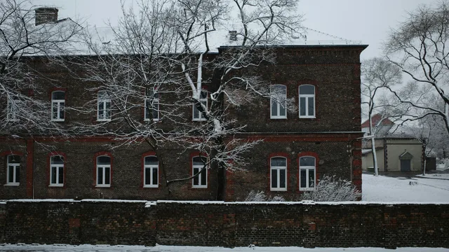 A long two-storey red-brick building with arched windows and a chimney on its snow-covered roof, bare trees in front of it in falling snow — the kind of older masonry building whose commercial boiler is started up again each autumn before the cold arrives