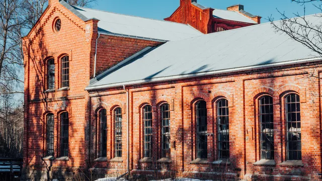 A long red-brick workshop with tall arched windows and a snow-covered roof, low winter sun across the brickwork — the kind of shop building whose exhaust and make-up air have to keep working through a cold season