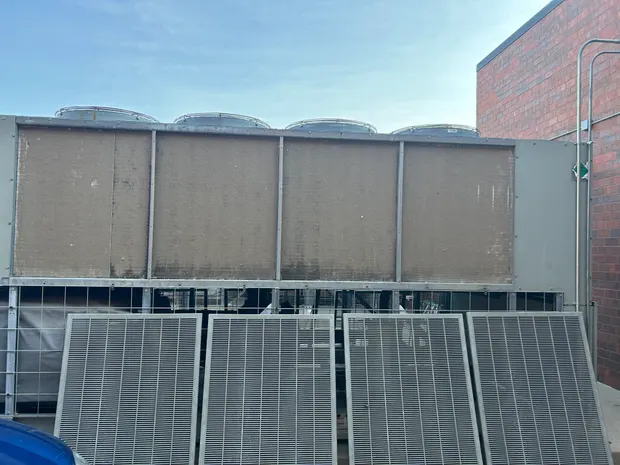 Commercial cooling tower with four top-mounted fans and weather-stained intake faces, its louvred panels set out on the ground in front of it