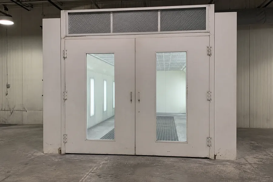 An enclosed spray paint booth standing in a shop bay, its two doors carrying tall viewing windows and a bank of intake filters across the top