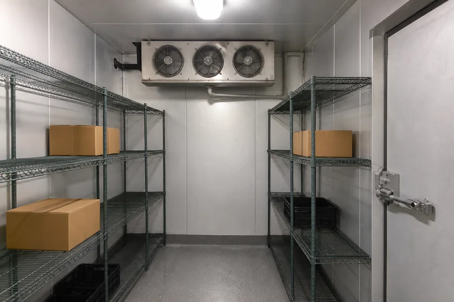 The interior of a walk-in cooler, a three-fan evaporator mounted high on the back wall above insulated panel walls, with wire shelving down both sides