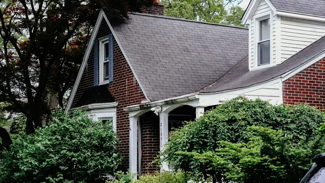 A brick house with a steep shingled roof and a dormer window, its front screened by overgrown shrubs