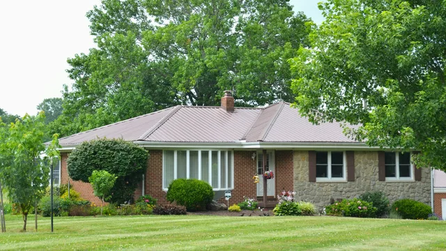 A single-storey brick and stone house with a metal hip roof, mature green trees behind it and planting beds along the front