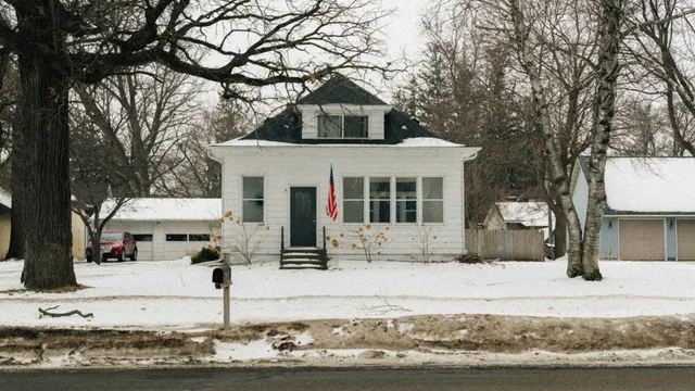 A white clapboard house with a dormer, set back from a road behind snow-covered ground and bare trees