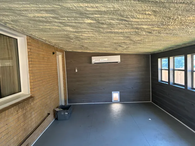 Ductless mini-split head mounted high on the plank wall of an enclosed porch, with a spray-foamed ceiling and the house’s original brick wall alongside