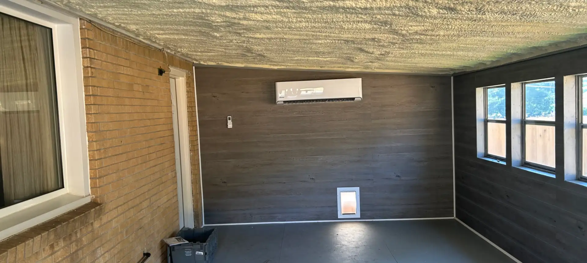 Ductless mini-split head mounted high on the plank wall of an enclosed porch, with a spray-foamed ceiling and the house’s original brick wall alongside