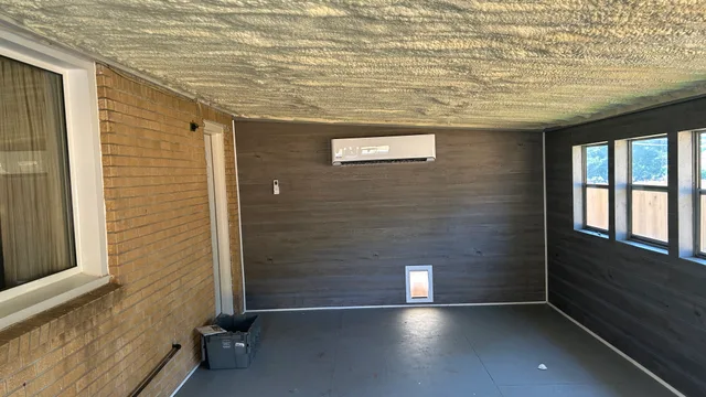 Ductless mini-split head mounted high on the plank wall of an enclosed porch, with a spray-foamed ceiling and the house’s original brick wall alongside