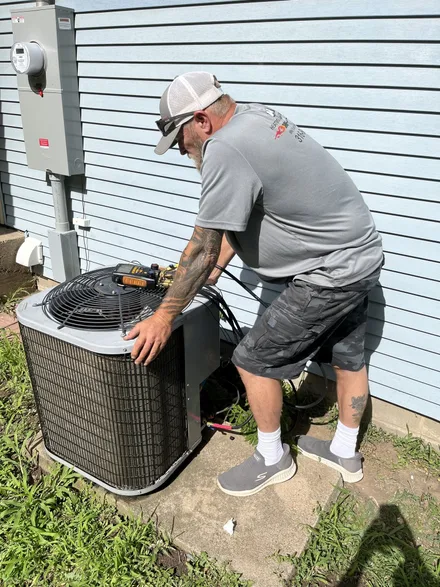 Jesse Mort, in an Arrow-branded shirt and cap, servicing an outdoor air conditioning condenser with manifold gauges set on top of the unit