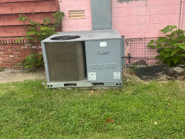 Weathered Armstrong Air packaged unit on rails against a painted block wall, with sheet-metal duct rising up the wall behind it