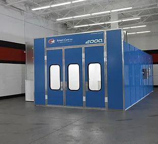 Enclosed blue spray paint booth with three viewing-window doors, standing on the concrete floor of a shop bay